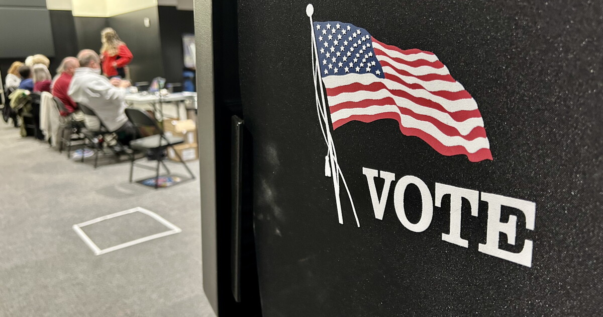 A black voting booth marked with a U.S. flag and “I VOTE” stands inside a polling location.